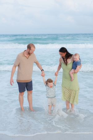 Happy family of four walking in ocean waves, parents holding toddler's hands, mom carrying baby.
