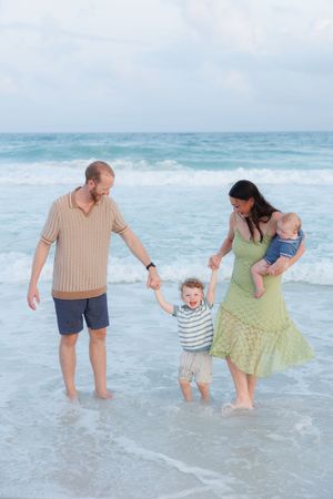 Family of four enjoying a beach day, parents holding toddler's hands in ocean waves while mother carries baby.