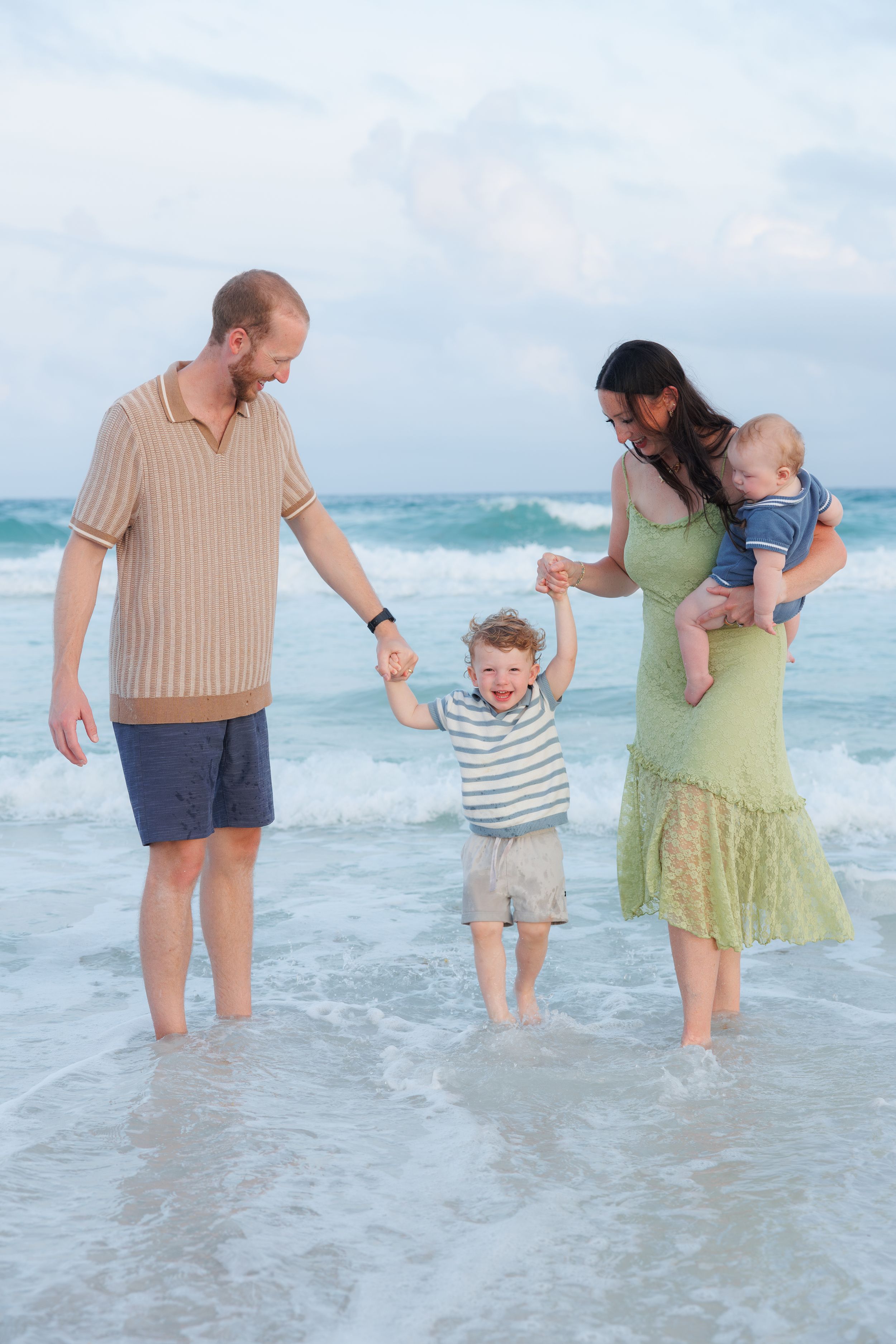 Happy family of four playing in shallow ocean waves on a cloudy beach day, mom holding infant and toddler.