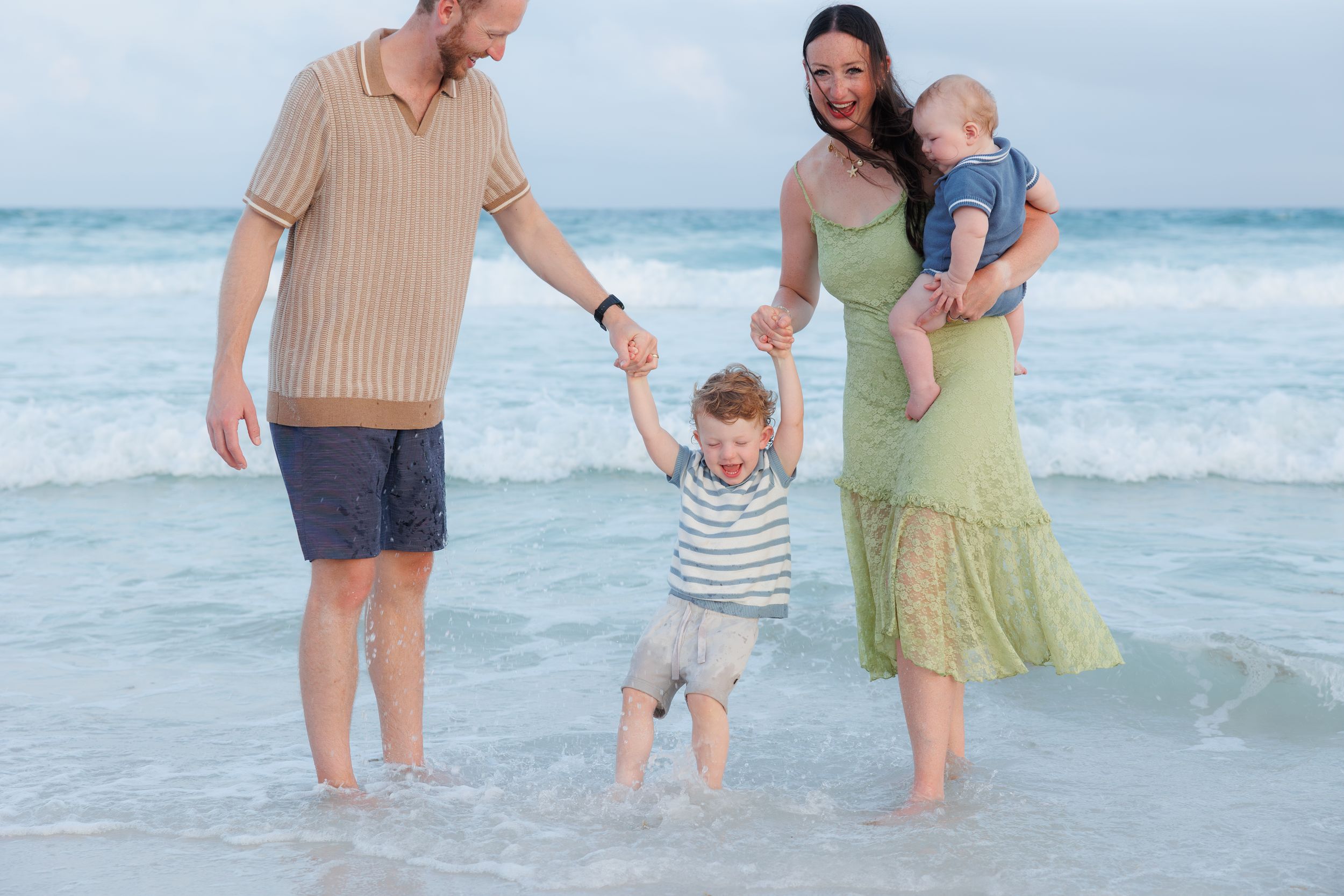 Family of four enjoying a beach day, parents holding toddler's hands in ocean waves, mother carrying baby.