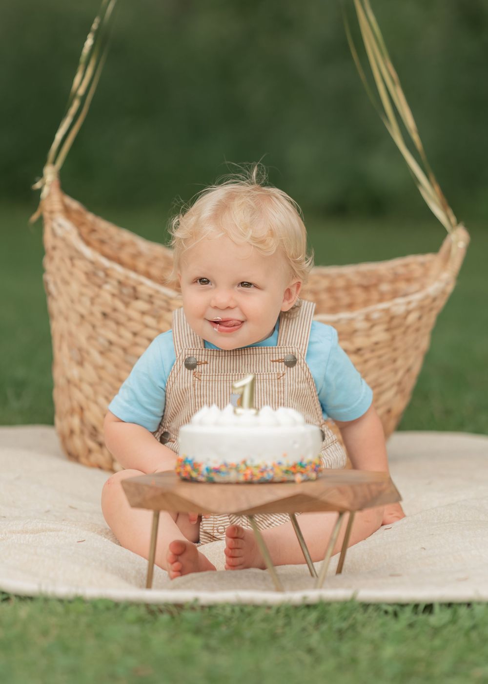 Happy toddler in tan overalls sits with a white sprinkle first birthday cake in a garden setting.