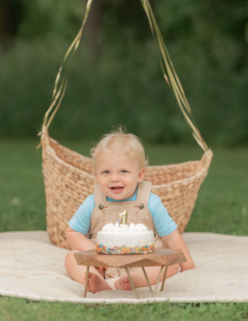 Smiling baby boy in tan overalls holds a first birthday cake with a number 1 candle outdoors.