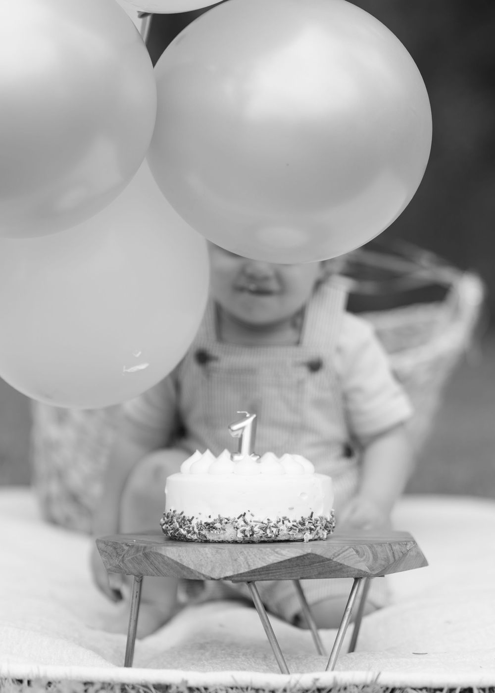 Black and white photo of a baby celebrating their first birthday with balloons and a cake with a number one candle.