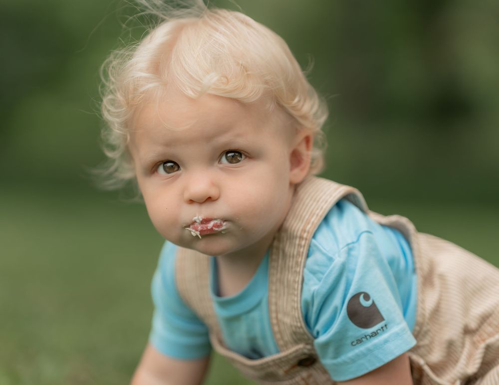 Curious blonde toddler in Carhartt overalls crawling outdoors with a flower in his mouth.