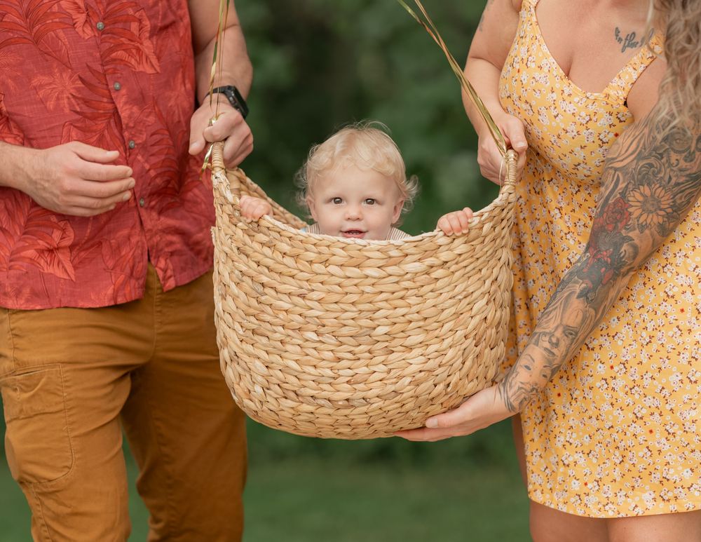 A toddler sits inside a woven basket held by two adults outdoors, one in a floral shirt and one in a yellow dress.