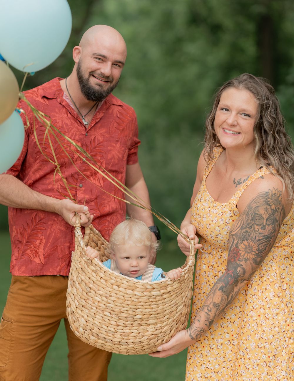 Parents hold baby in wicker basket with pastel balloons at outdoor first birthday celebration.