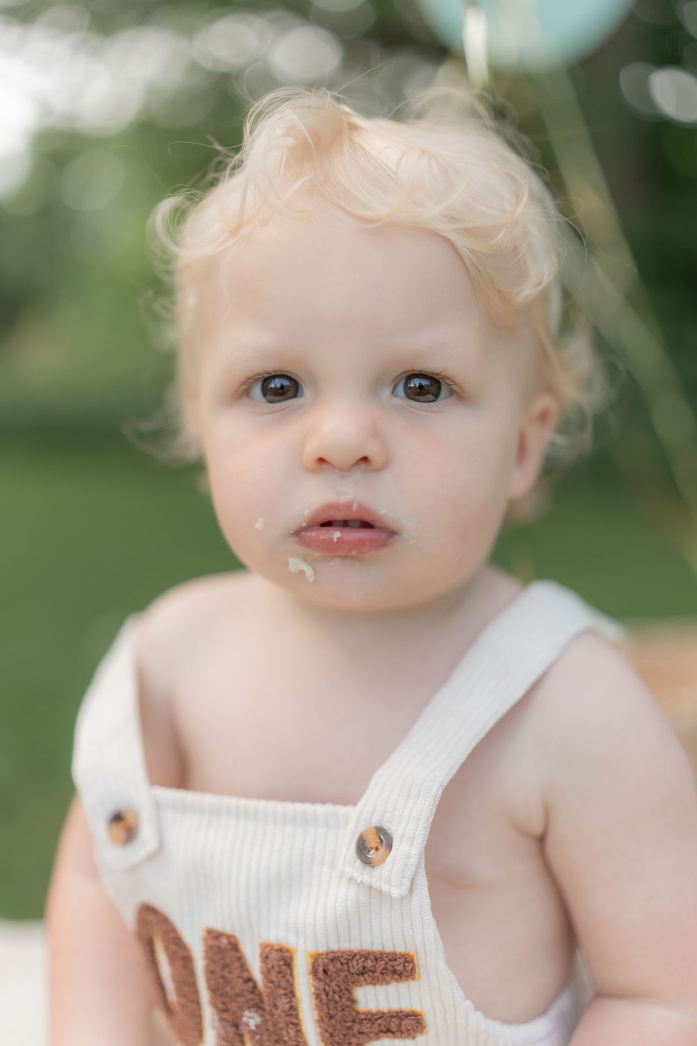 Close-up portrait of a blonde toddler in white ONE overalls with frosting on their face outdoors.