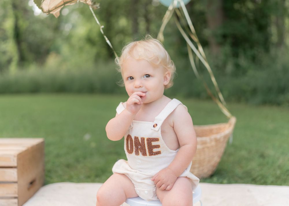 Toddler in white ONE romper sits outdoors for first birthday cake smash photo session.