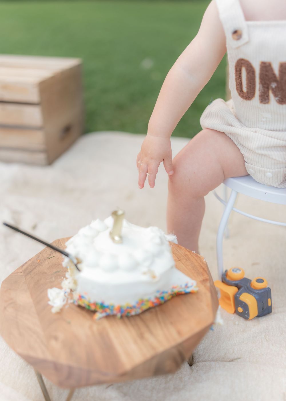Baby in ONE romper reaching toward smashed first birthday cake with sprinkles on a wooden stand outdoors.