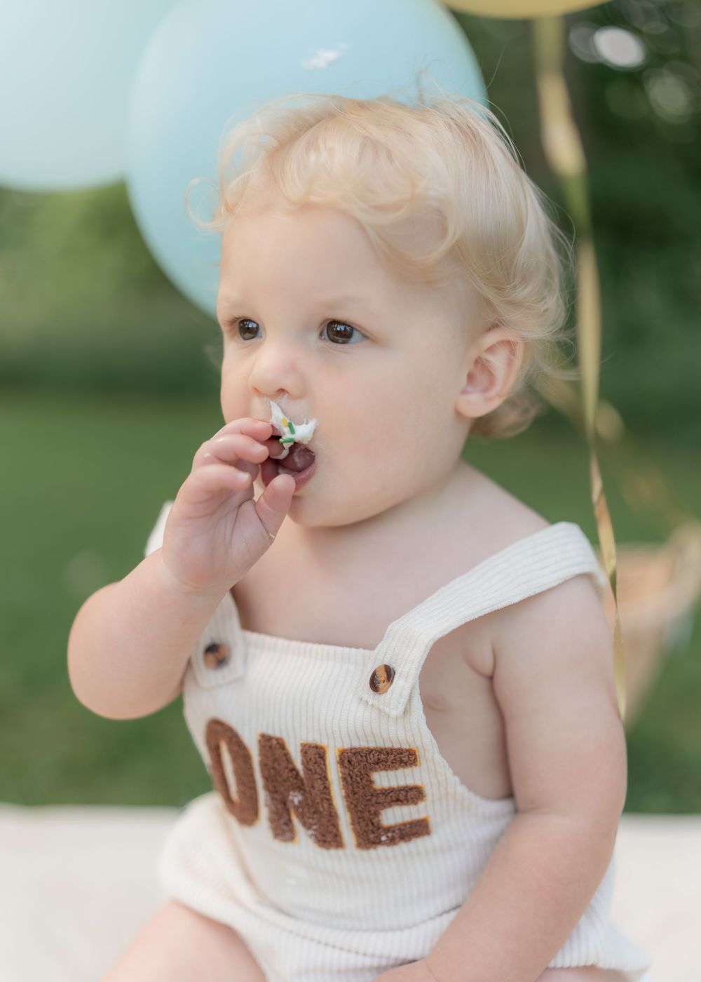 Blonde baby in ONE romper eats flower decoration at outdoor first birthday photo session.