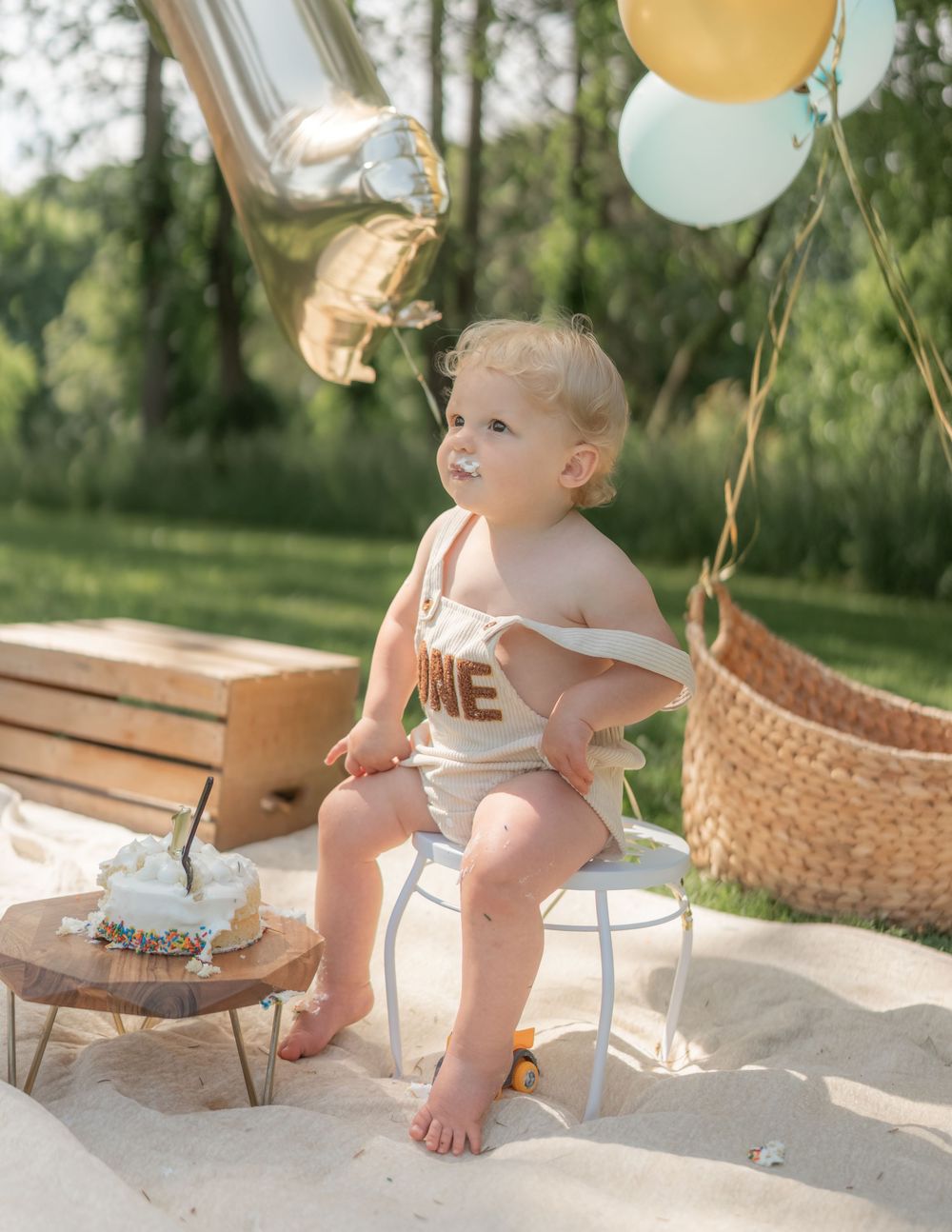 Baby in ONE romper sits on chair with smash cake and balloons at outdoor first birthday party.