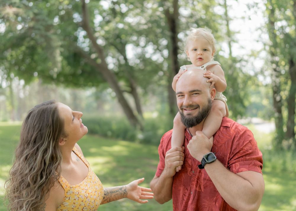Happy family outdoors, bald father holding toddler on shoulders while mother smiles in sunny green park.