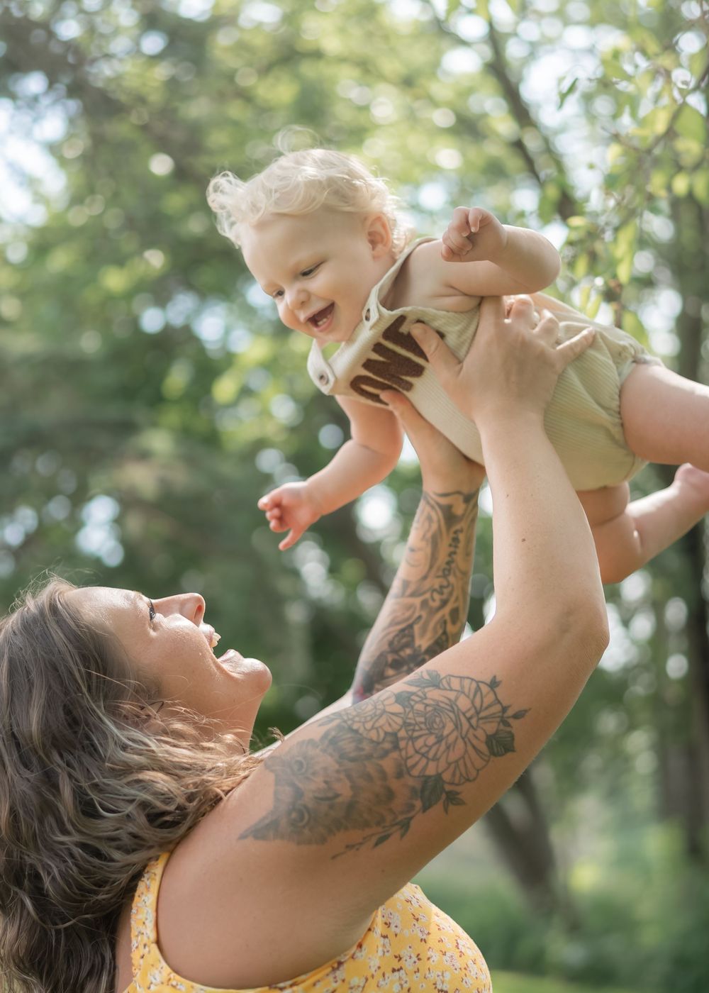 Tattooed mother joyfully lifting laughing baby in the air outdoors among green trees.