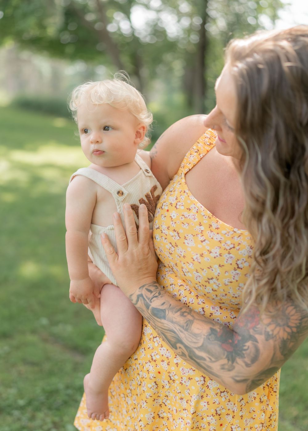 Tattooed mother in yellow floral dress holding blonde baby boy in a park setting.