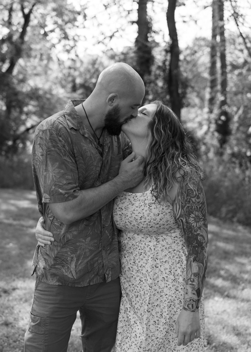 Black and white photo of a bald bearded man kissing a curly-haired tattooed woman outdoors.