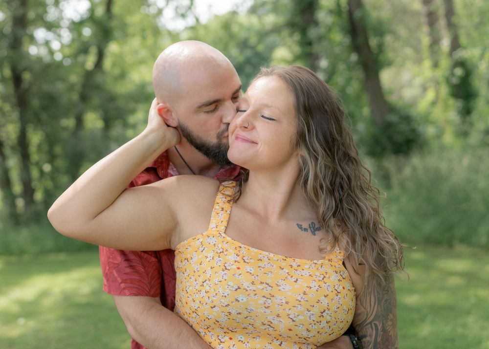A bald man in a red shirt kisses a smiling woman in a yellow floral dress outdoors in a lush green park.