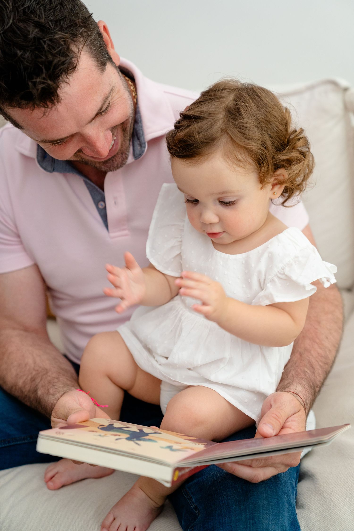 Father in pink shirt reads colorful picture book with toddler daughter in white dress on couch.