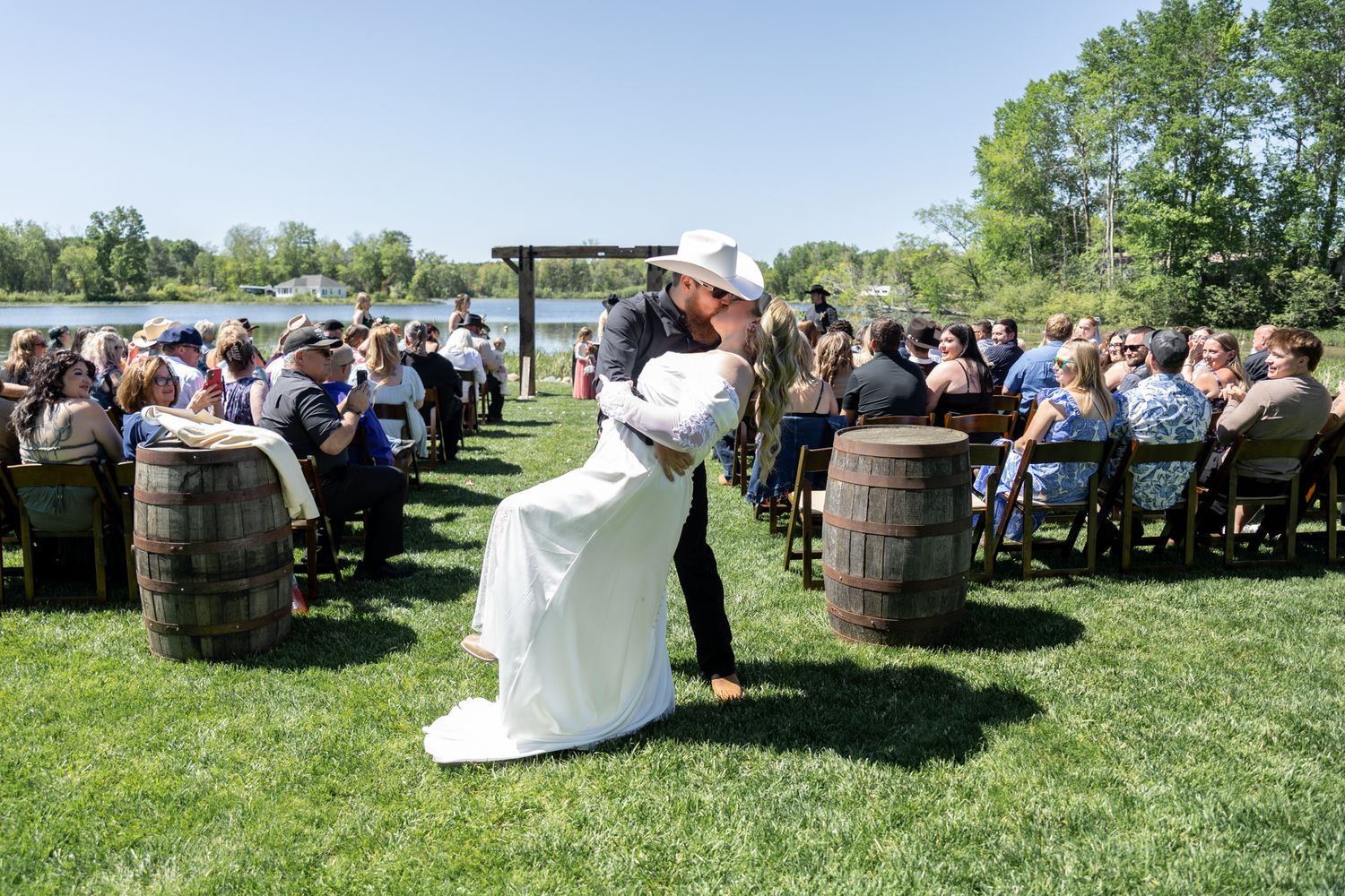 Bride and groom share a romantic embrace at an outdoor wedding ceremony by a river, flanked by rustic wooden barrels.
