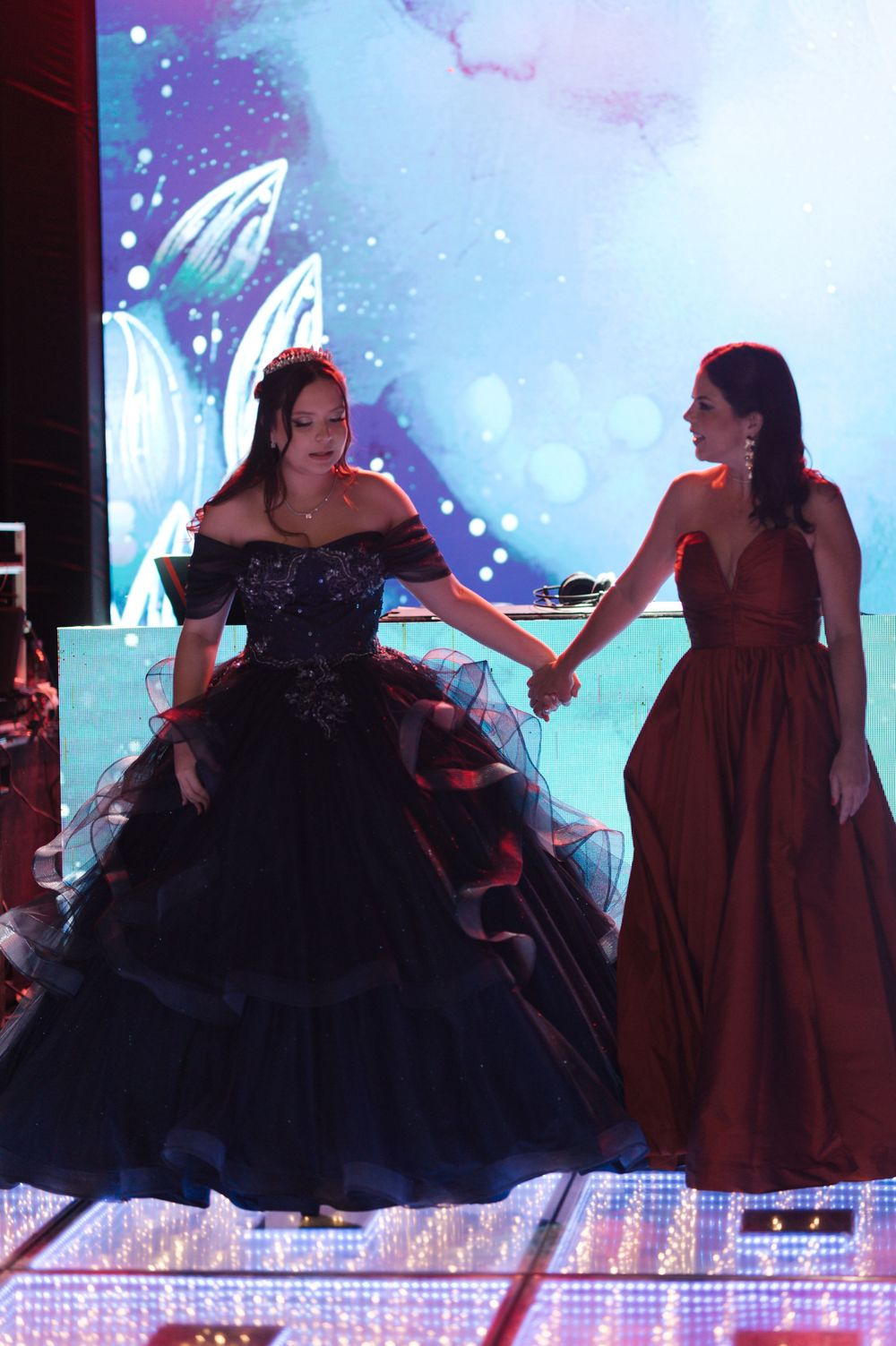 Two performers in contrasting black and burgundy formal dresses dance together on stage with a sparkly backdrop.
