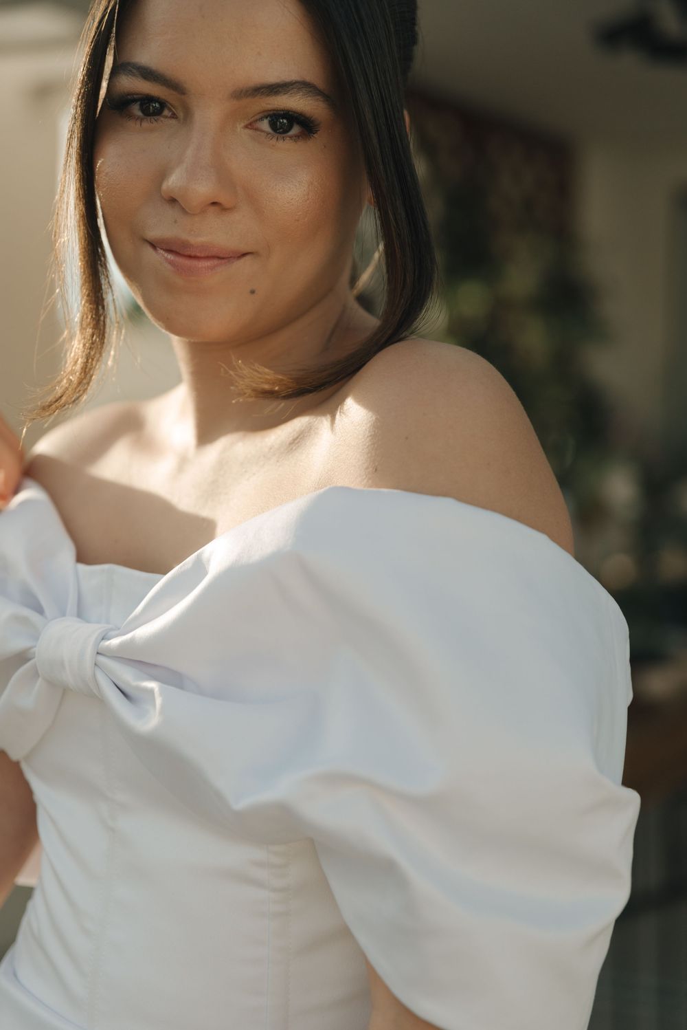 A person wearing an elegant white off-shoulder dress with bow detail poses in soft natural lighting.