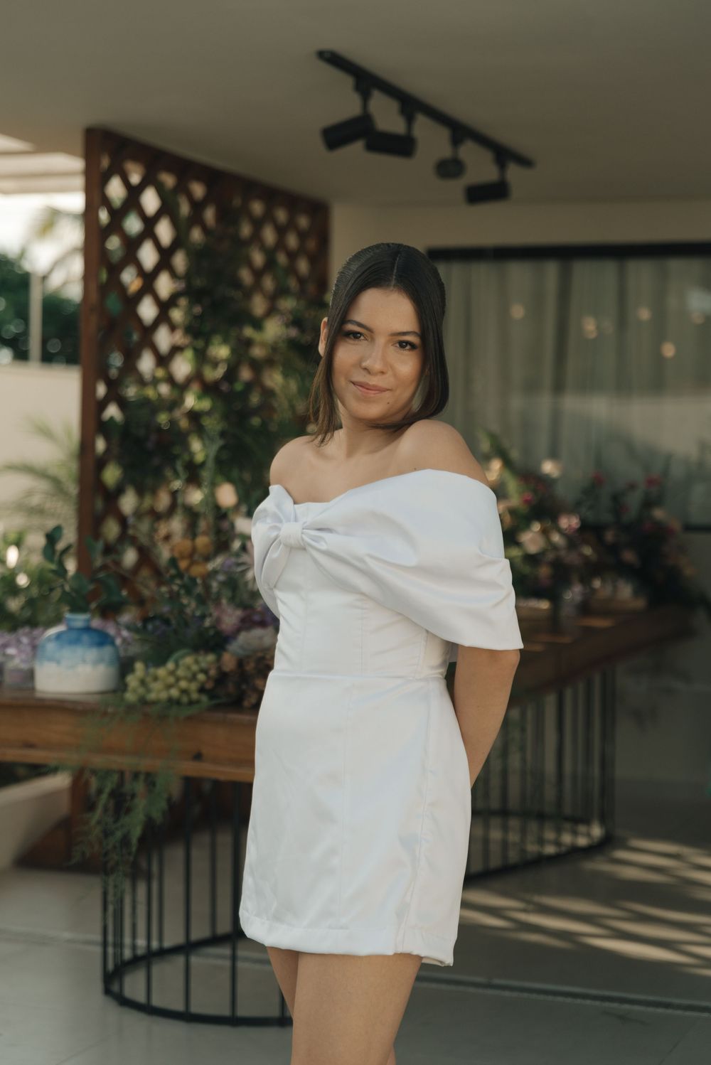 A white off-shoulder mini dress is shown in an indoor setting with decorative lattice and plants in the background.