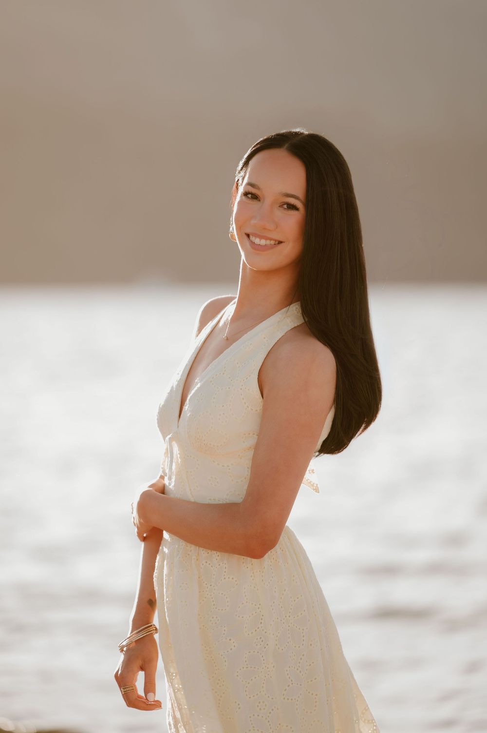 Smiling young woman with dark hair wearing a white dress poses gracefully near a sunlit waterfront.