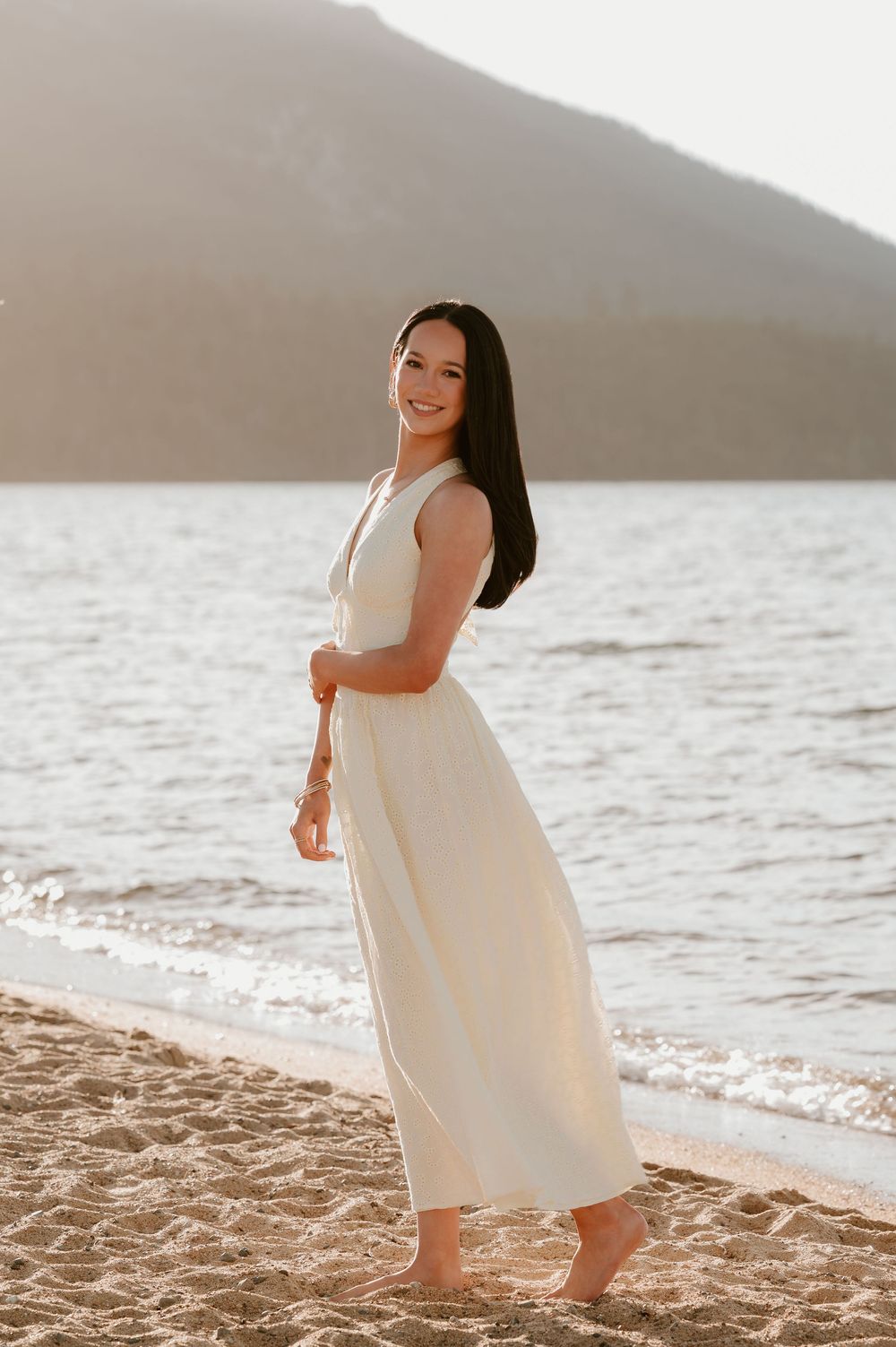 Young woman in white maxi dress smiling on sandy beach at golden hour with mountain and lake backdrop.