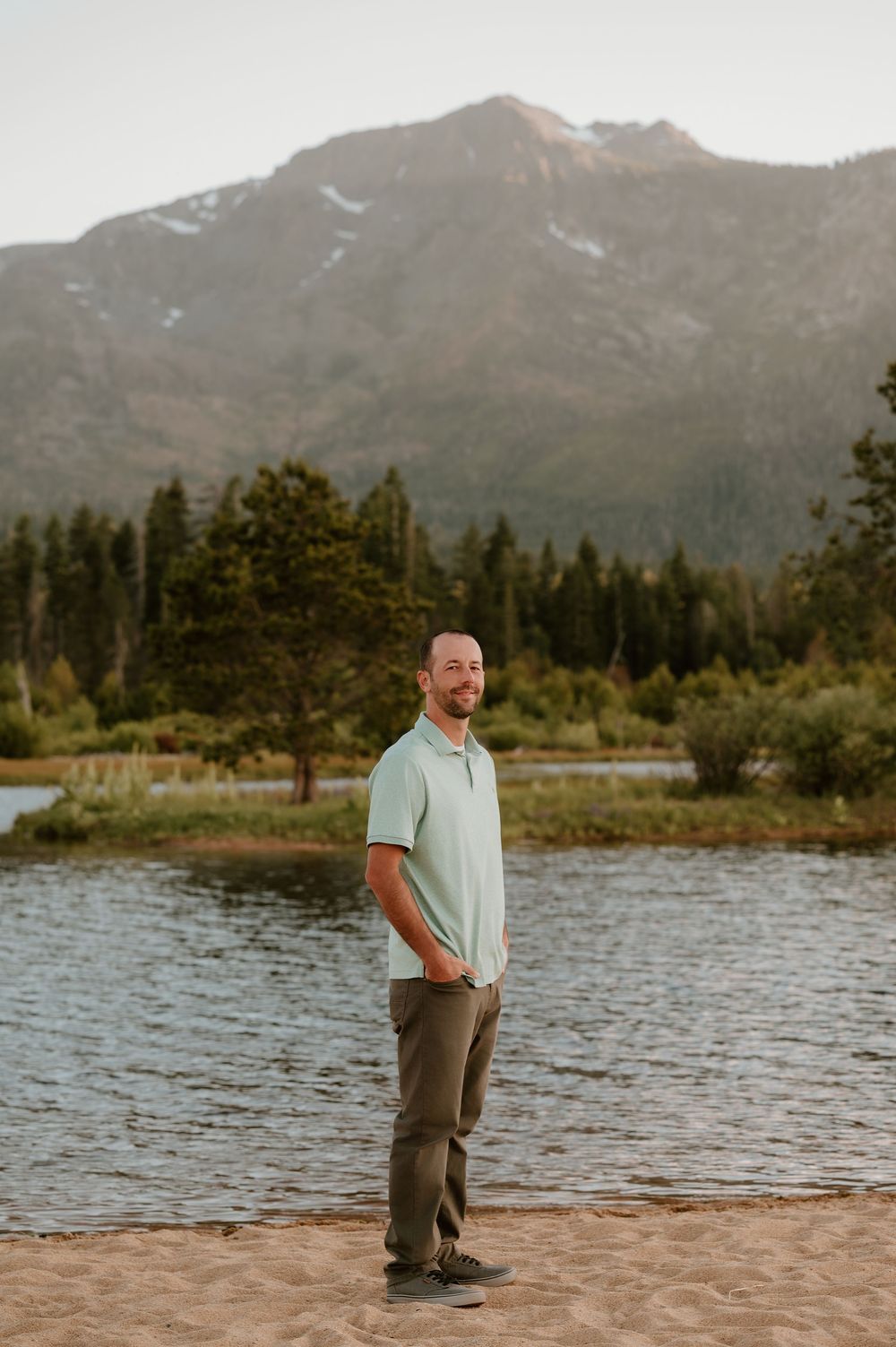 Man in mint polo and olive pants stands on sandy shore with mountain lake and pine trees in background.