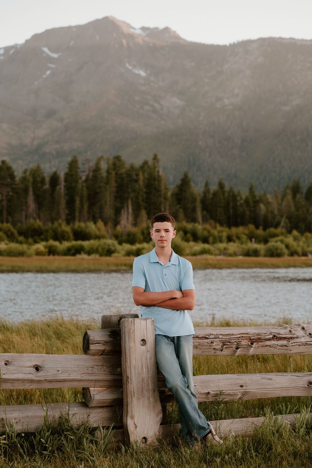 Teen boy in light blue polo leans on wooden fence with mountain lake and pine forest backdrop.