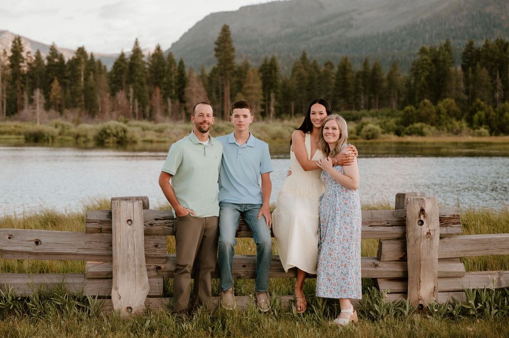 Family of four posing by a rustic wooden fence with a mountain lake and forest backdrop.