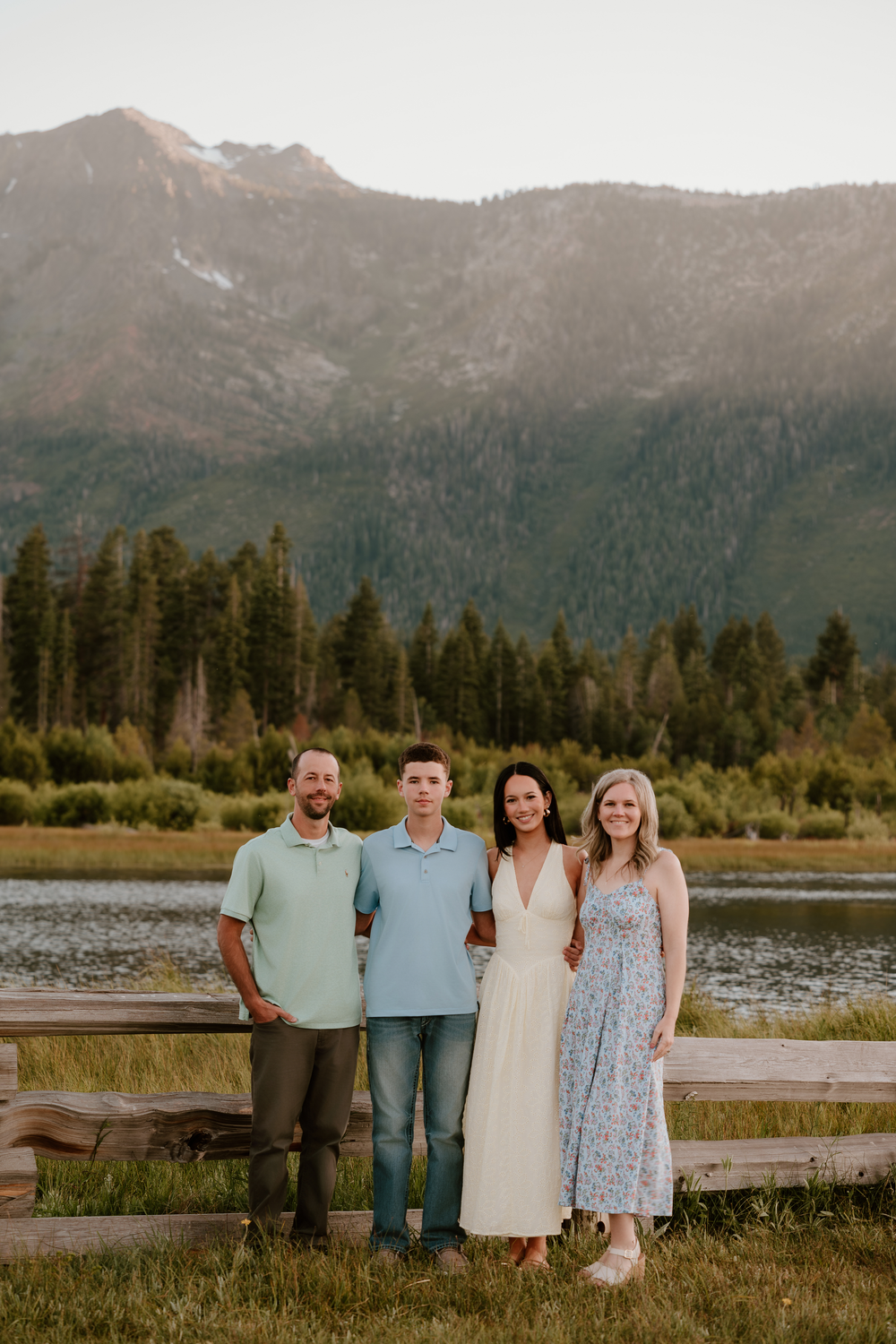 Family of four smiling together near a mountain lake with pine trees and peaks in the background.