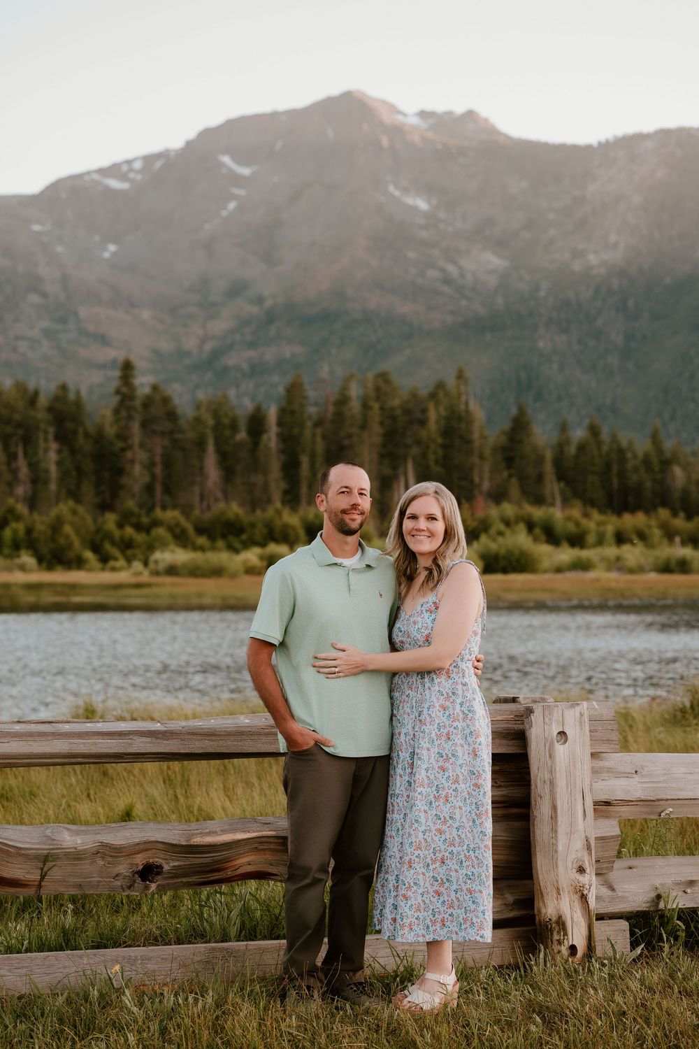 Couple posing by a wooden fence with a mountain lake and pine forest backdrop during golden hour.