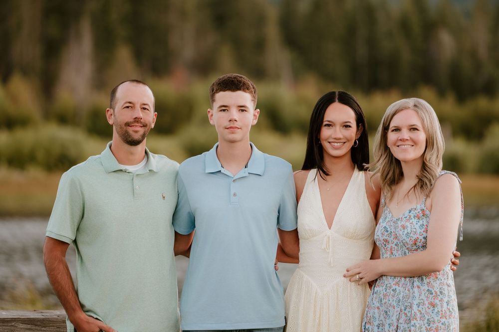 A smiling family of four poses outdoors near a river with mountains and trees in the background.