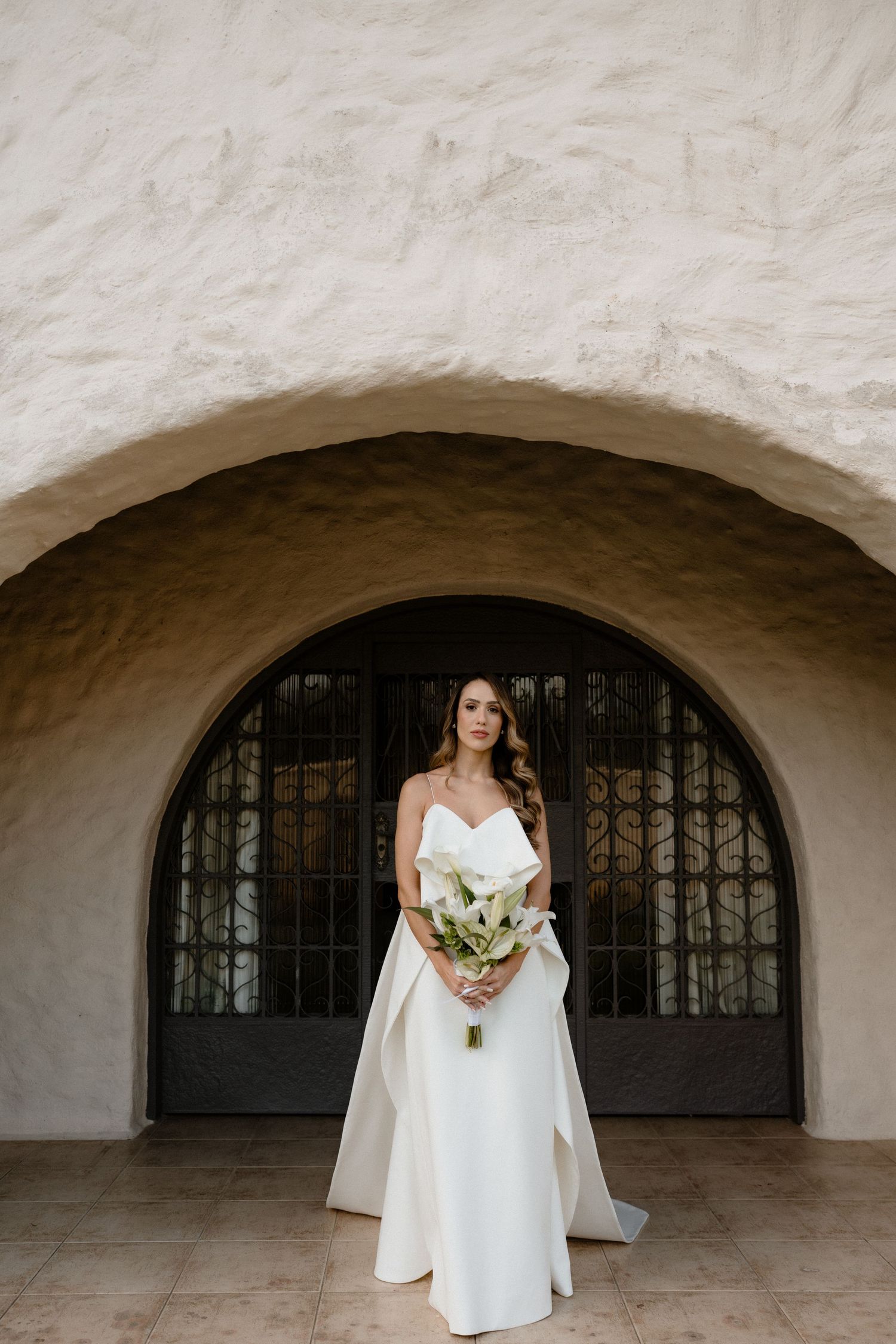 Bride in white gown holds bouquet under stone arch doorway, with small dog in gray outfit beside her.