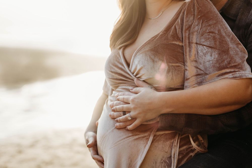 Pregnant woman in beige velvet dress being embraced by partner outdoors in soft golden light.
