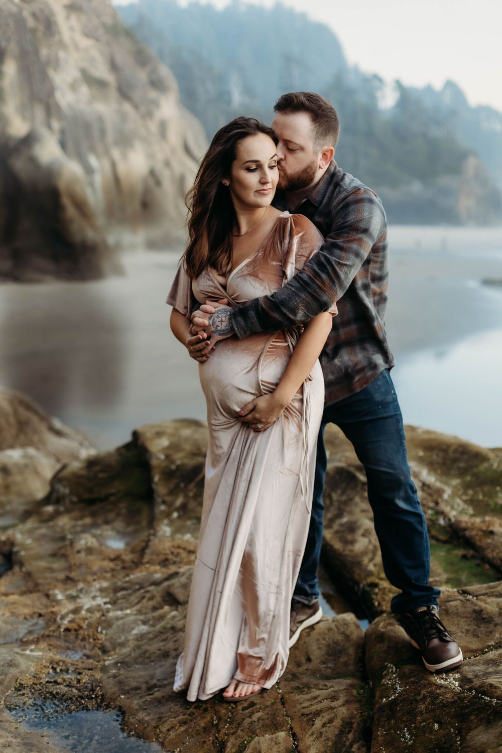 Couple embracing on rocky shore, man kissing pregnant woman in blush gown, misty cliffs and lake backdrop.