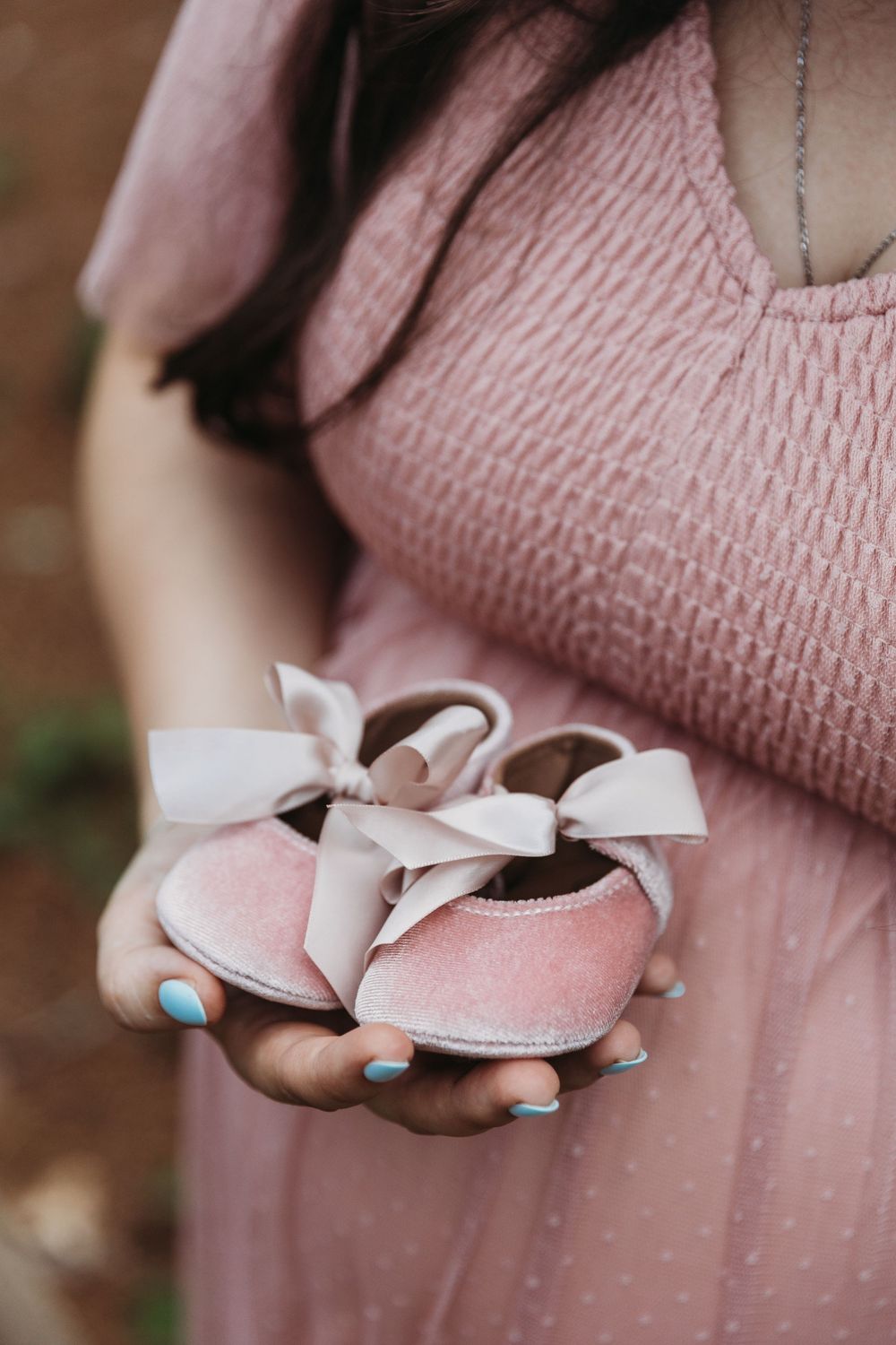Pregnant woman in pink dress holding tiny pink velvet baby shoes with white bows, announcing a baby girl.