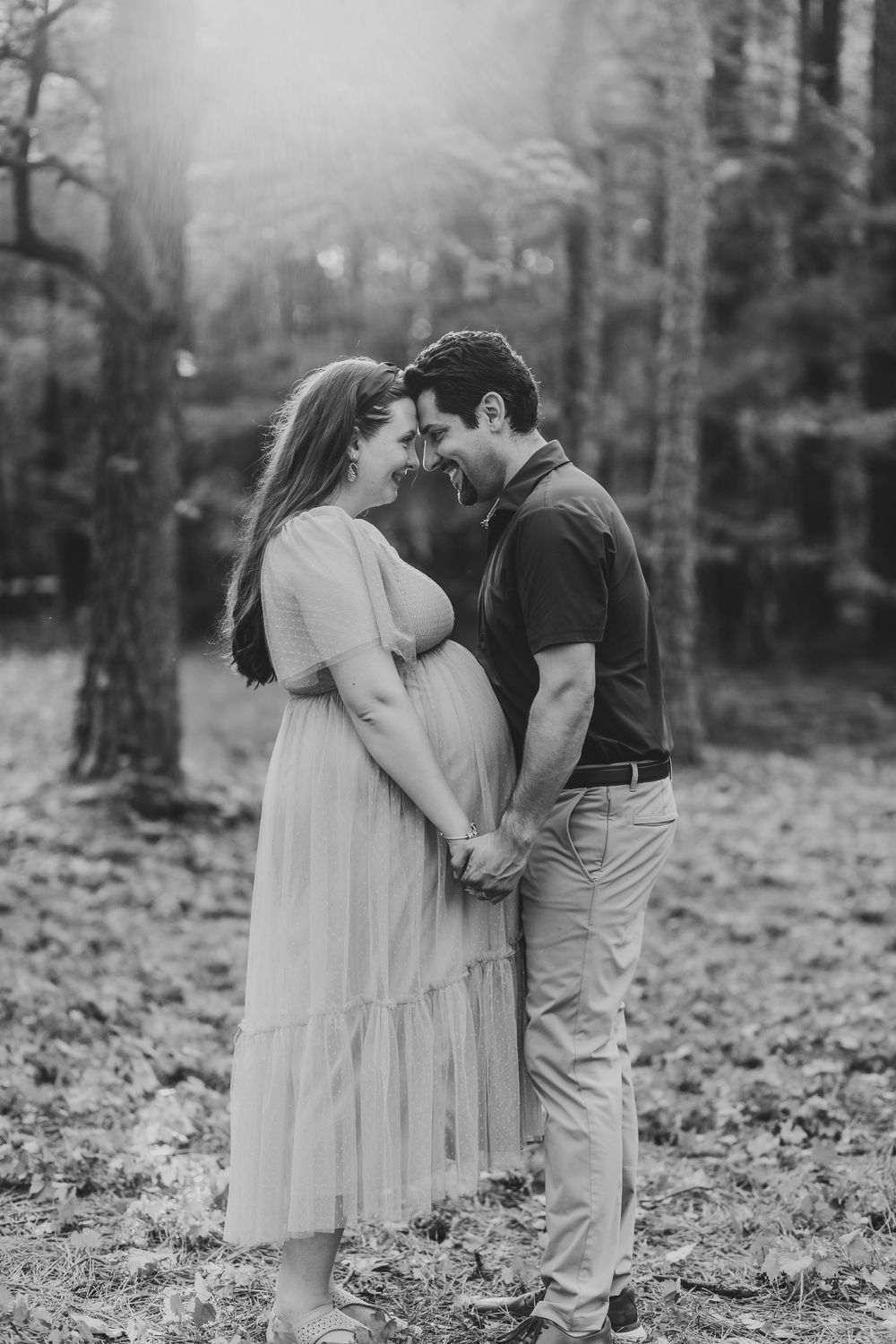 Black and white maternity photo of couple holding hands and touching foreheads in a forest setting.