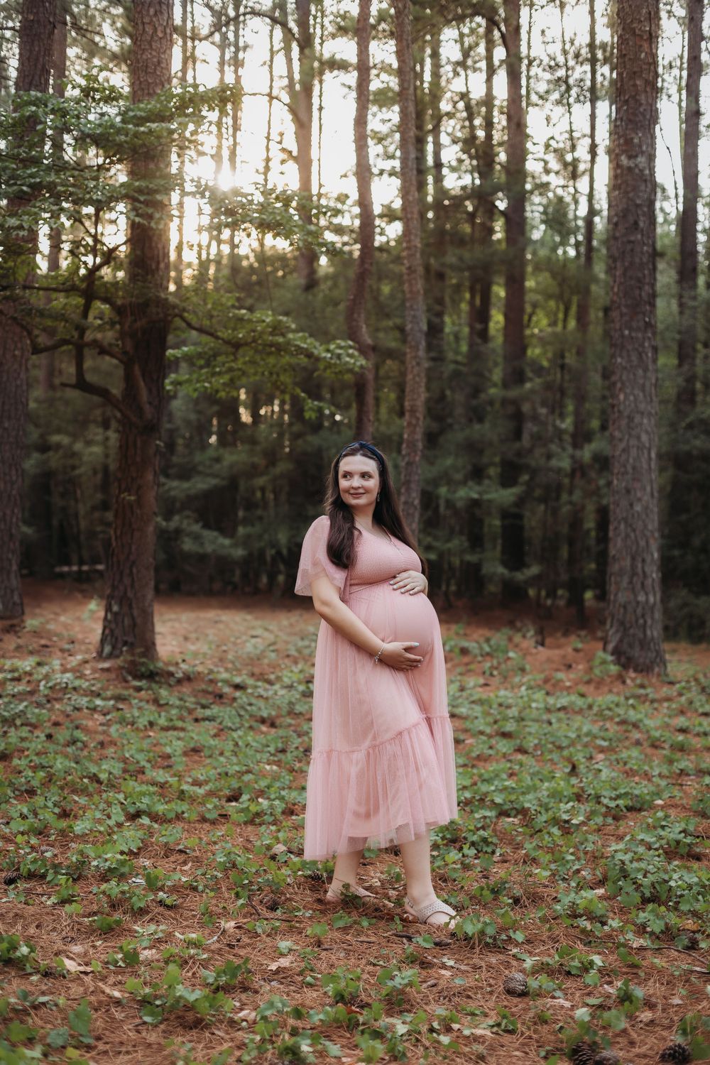Pregnant woman in pink tulle dress cradling baby bump while standing in a sunlit pine forest.