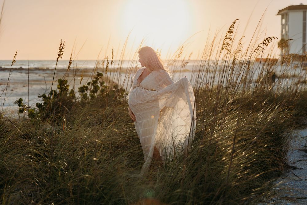 Pregnant woman in flowing white dress standing in beach dunes at golden sunset with sea oats and ocean backdrop.