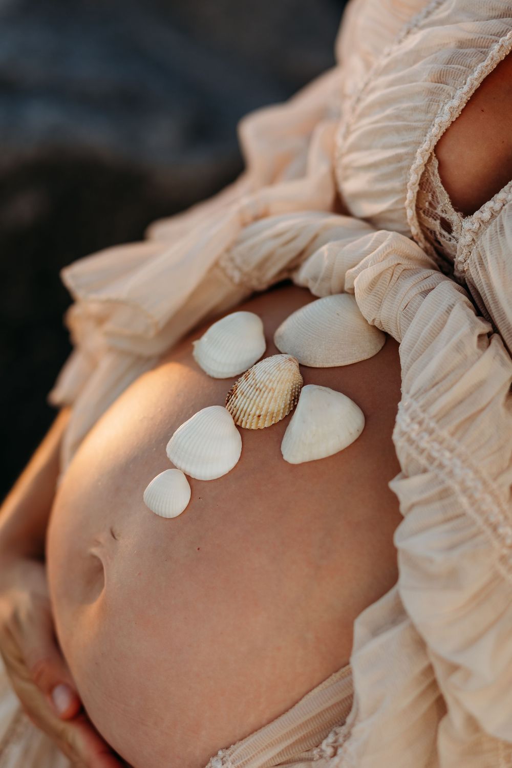 Pregnant woman in beige dress with seashells arranged on her baby bump, captured in warm golden light.