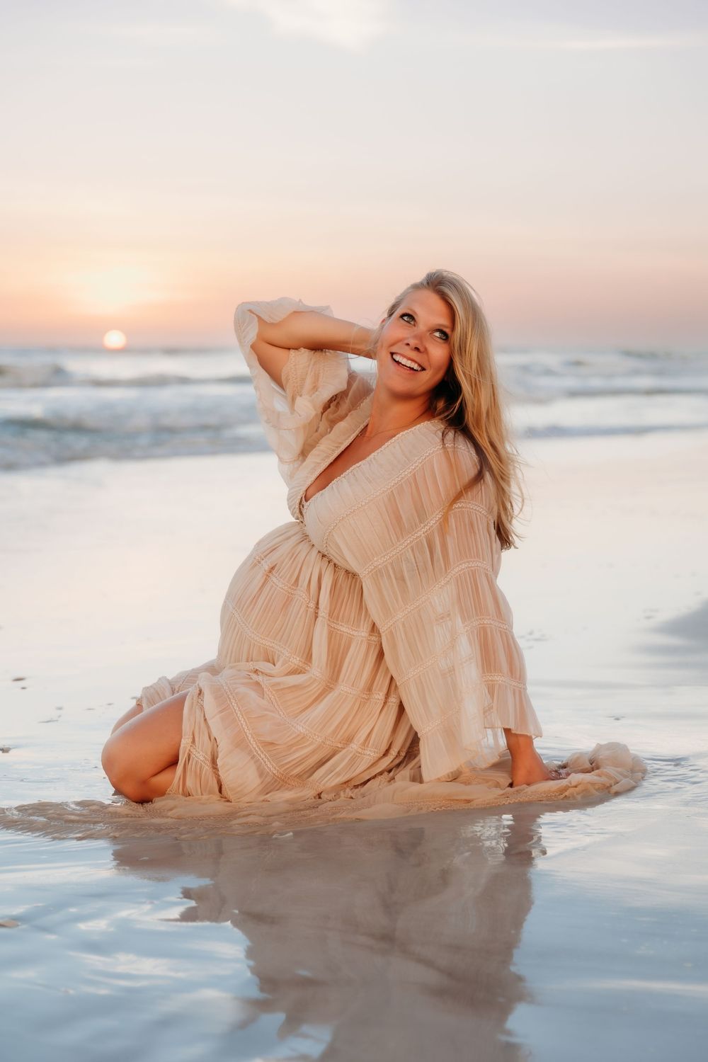 Smiling pregnant woman in beige dress sitting on sandy beach at sunset with ocean waves.
