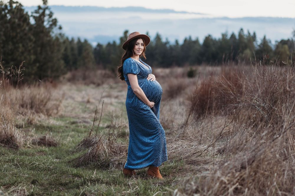 Pregnant woman in blue maxi dress and hat poses in a field with trees in the background.