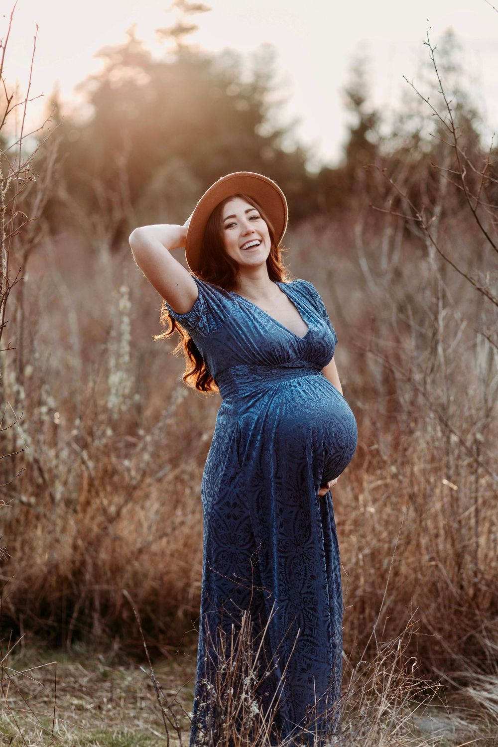 Smiling pregnant woman in blue dress and brown hat standing in golden field at sunset.