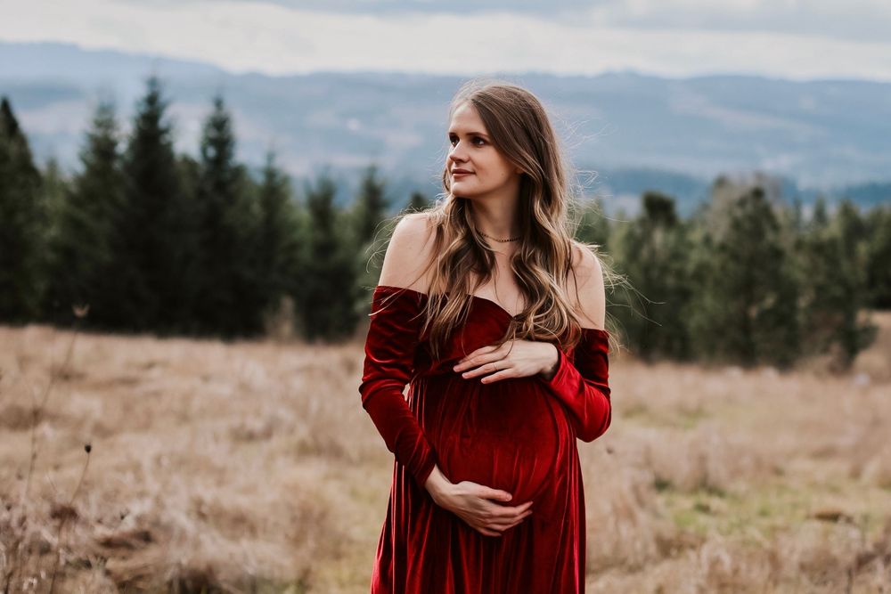 Pregnant woman in red velvet off-shoulder gown cradling belly in scenic mountain meadow with evergreen trees.