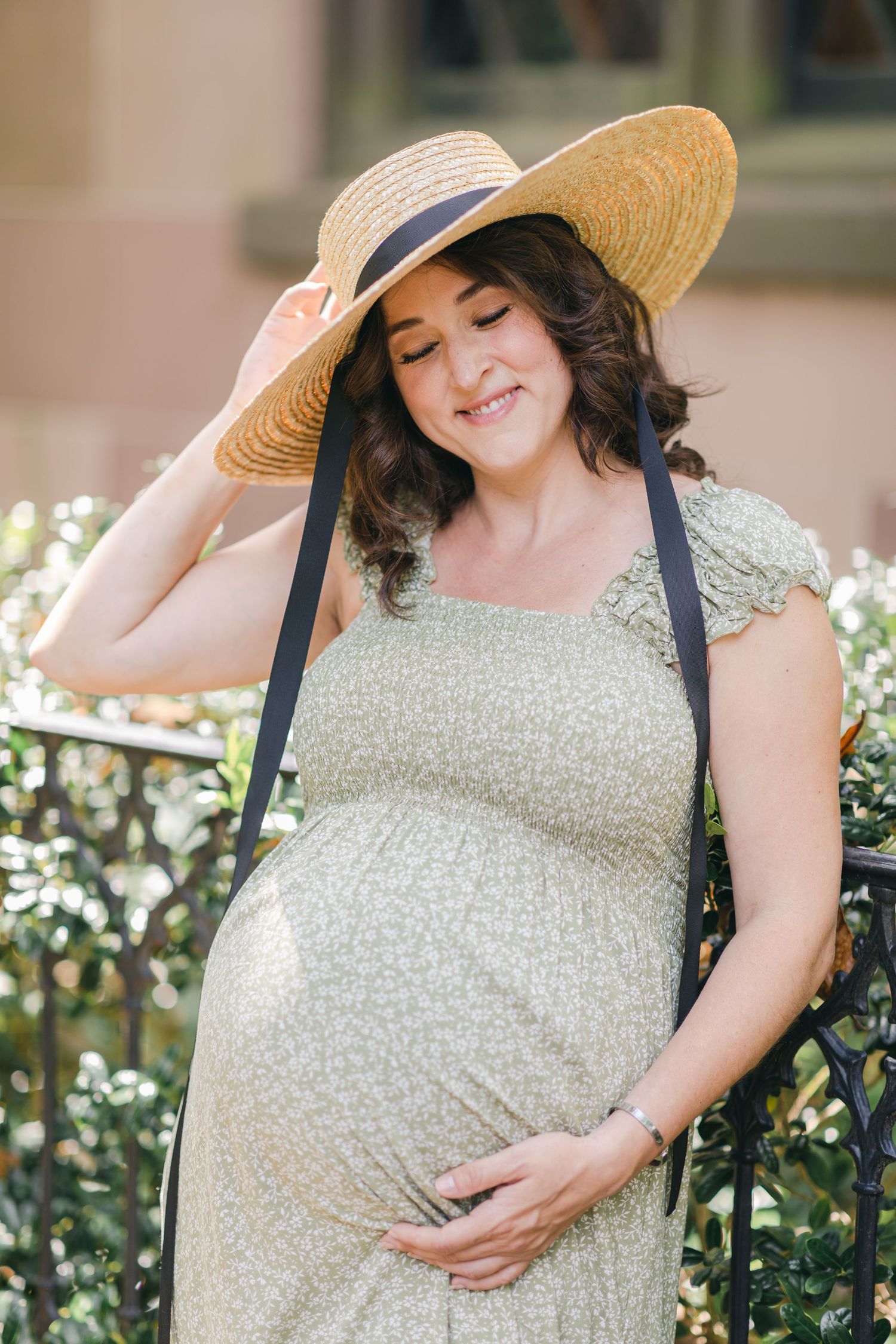 Pregnant woman in floral dress and straw hat smiling while cradling her baby bump outdoors.
