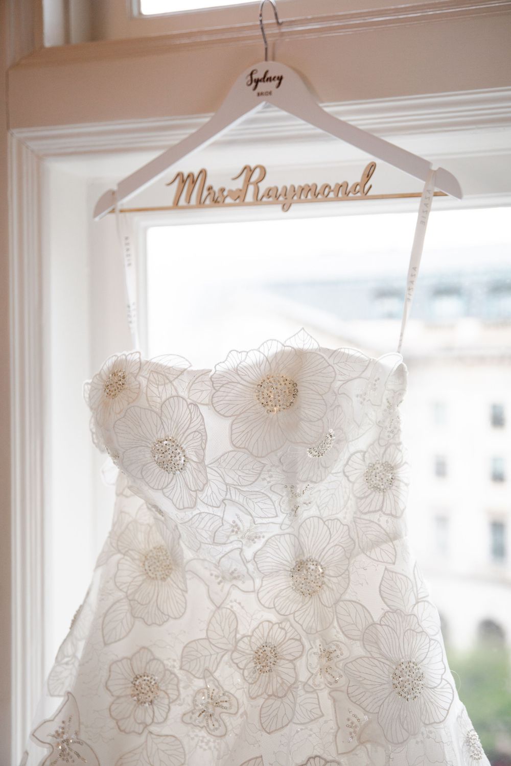 A strapless floral lace wedding dress hanging on a personalized Mrs. Raymond hanger in a bright window.