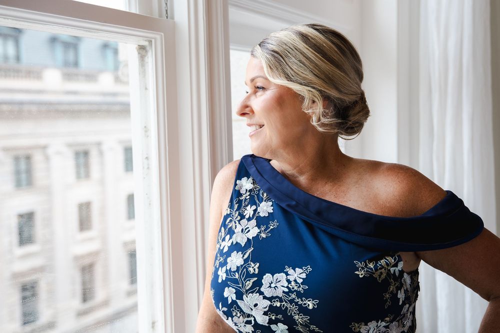 Elegant blonde woman in blue floral off-shoulder dress gazing thoughtfully out a window in a bright room.