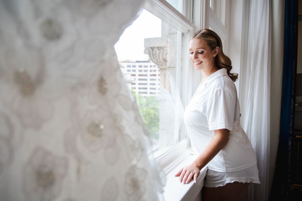 Bride in white robe sitting by window with wedding dress displayed, smiling softly in elegant hotel room.