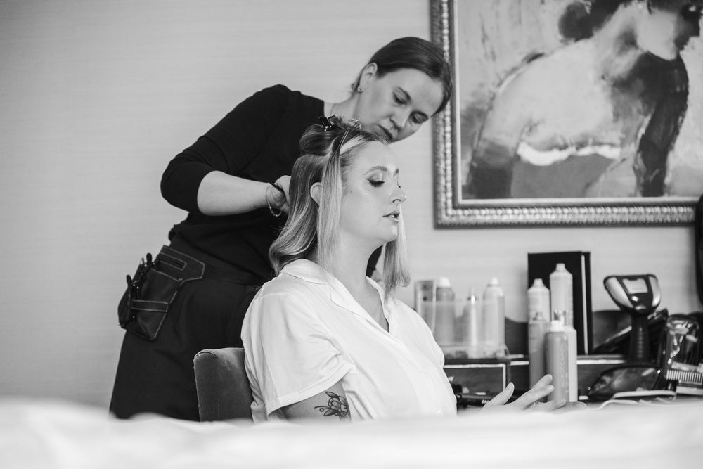 Black and white photo of a hairstylist styling a seated blonde woman's hair in a bright salon setting.